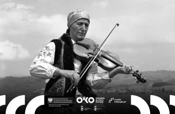 Starsza kobieta w tradycyjnym stroju ludowym siedzi na drewnianej ławce i gra na skrzypcach, w tle widać rozległy krajobraz wiejski.