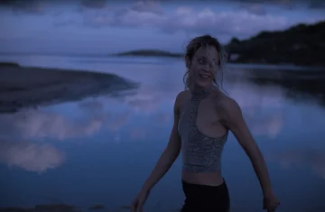 Kobieta stojąca przy spokojnej wodzie o zmierzchu, ubrana w sportowy top, z rozwianymi włosami, patrząca w bok; w tle odbicie nieba i ciemny krajobraz.