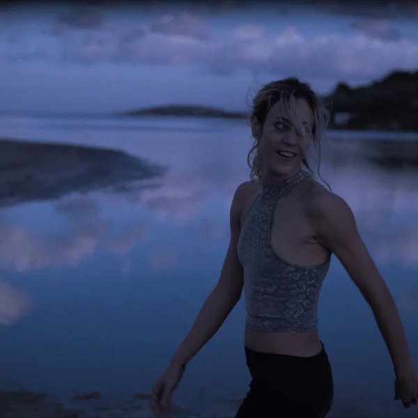 Kobieta stojąca przy spokojnej wodzie o zmierzchu, ubrana w sportowy top, z rozwianymi włosami, patrząca w bok; w tle odbicie nieba i ciemny krajobraz.