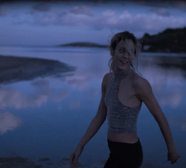 Kobieta stojąca przy spokojnej wodzie o zmierzchu, ubrana w sportowy top, z rozwianymi włosami, patrząca w bok; w tle odbicie nieba i ciemny krajobraz.