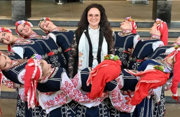 zdjęcie przedstawia kobietę w długich włosach otoczoną kobietami w strojach ludowych z Bułgarii