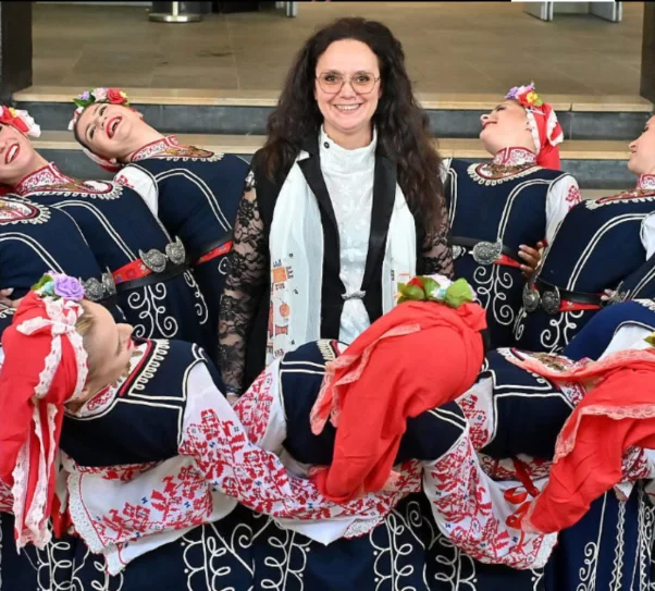 zdjęcie przedstawia kobietę w długich włosach otoczoną kobietami w strojach ludowych z Bułgarii