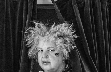 Czarno-białe zdjęcie przedstawia kobietę stojącą między ciemnymi, ciężkimi zasłonami. Nad jej głową widoczny jest znak z napisem „WYJŚCIE EWAKUACYJNE”. Kobieta ma kręcone, potargane włosy i wyrazisty makijaż z ozdobnym wzorem pod okiem.