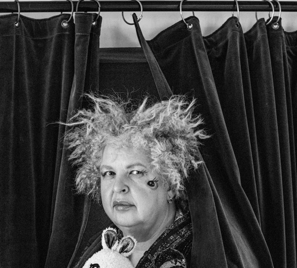 Czarno-białe zdjęcie przedstawia kobietę stojącą między ciemnymi, ciężkimi zasłonami. Nad jej głową widoczny jest znak z napisem „WYJŚCIE EWAKUACYJNE”. Kobieta ma kręcone, potargane włosy i wyrazisty makijaż z ozdobnym wzorem pod okiem.