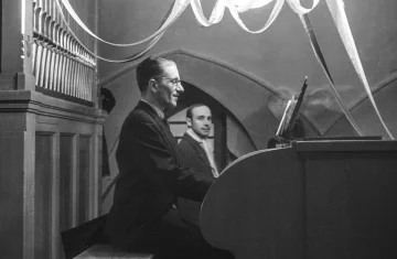 Czarno-białe zdjęcie przedstawiające mężczyznę siedzącego przy organach piszczałkowych w kościelnym wnętrzu. Gra na instrumencie, ubrany w ciemny garnitur. W tle widoczny jest drugi mężczyzna oraz dekoracje zawieszone pod sklepieniem.