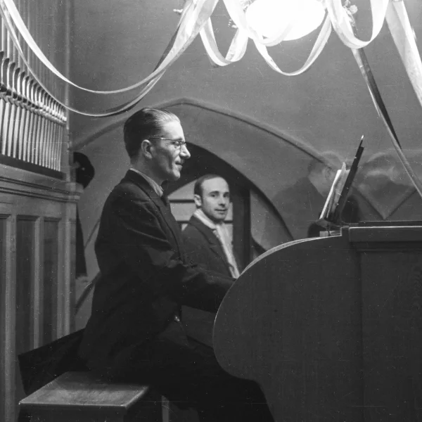 Czarno-białe zdjęcie przedstawiające mężczyznę siedzącego przy organach piszczałkowych w kościelnym wnętrzu. Gra na instrumencie, ubrany w ciemny garnitur. W tle widoczny jest drugi mężczyzna oraz dekoracje zawieszone pod sklepieniem.