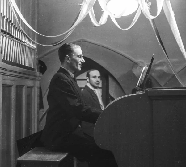 Czarno-białe zdjęcie przedstawiające mężczyznę siedzącego przy organach piszczałkowych w kościelnym wnętrzu. Gra na instrumencie, ubrany w ciemny garnitur. W tle widoczny jest drugi mężczyzna oraz dekoracje zawieszone pod sklepieniem.