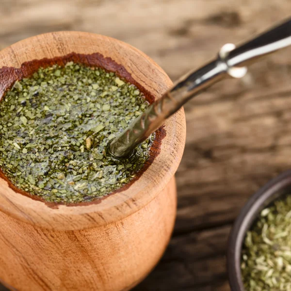 Drewniane naczynie z yerba mate wypełnione zielonym naparem z liści, z metalową rurką do picia zwaną bombillą, stojące na drewnianym stole.