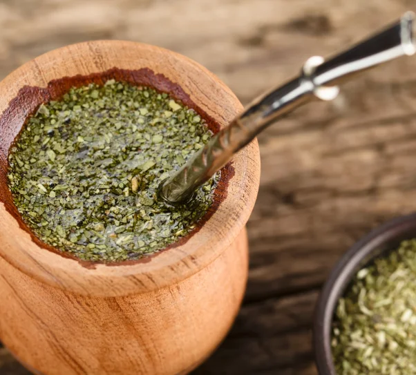 A wooden gourd filled with green yerba mate infusion and a metal straw called a bombilla, placed on a wooden table.