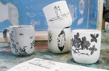 Cztery ceramiczne kubki ozdobione czarno-białymi grafikami stoją na stole. Na kubkach widać różne motywy: mrówki, liście, osobę na desce SUP oraz gałązkę z kwiatami. W tle znajduje się niebieskie sito drukarskie i fragment papieru z rysunkami technicznymi.