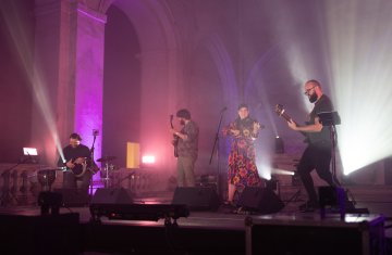 Na scenie jest zespół, dwóch mężczyzn gra na gitarach, trzeci mężczyzna siedzi przy bębnach, kobieta w sukience w kwiaty gra na skrzypcach