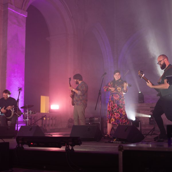 Na scenie jest zespół, dwóch mężczyzn gra na gitarach, trzeci mężczyzna siedzi przy bębnach, kobieta w sukience w kwiaty gra na skrzypcach