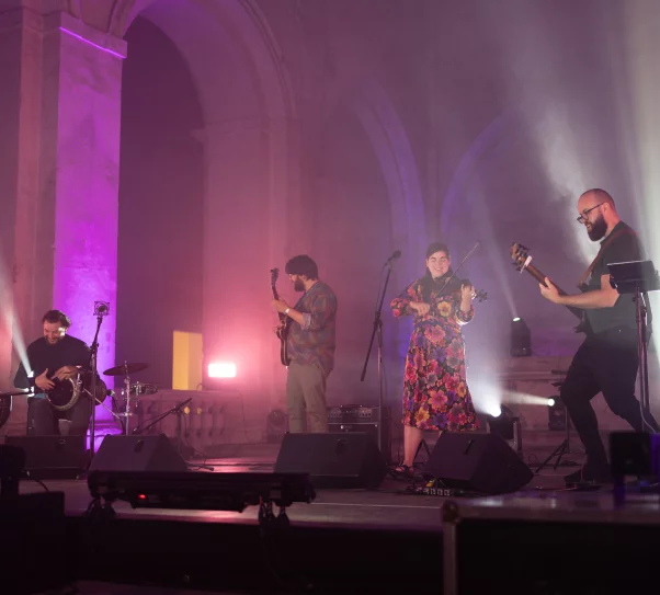 Na scenie jest zespół, dwóch mężczyzn gra na gitarach, trzeci mężczyzna siedzi przy bębnach, kobieta w sukience w kwiaty gra na skrzypcach