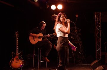 dziewczyna w białej bluzce śpiewa na scenie, w tle widać gitarzystę.