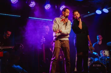 Na scenie dwoje wokalistów śpiewa do mikrofonów, stojąc obok siebie w kolorowych światłach reflektorów. W tle widać muzyków grających na gitarze i perkusji. Atmosferę występu podkreśla fioletowy dym sceniczny.