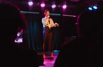 Zdjęcie przedstawia osobę stojącą na scenie i mówiącą do mikrofonu podczas występu. Scena jest oświetlona kolorowymi reflektorami, a na pierwszym planie widoczne są sylwetki publiczności.