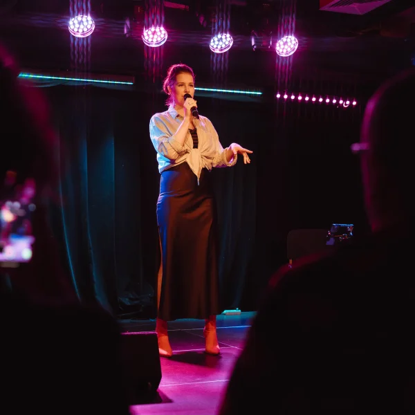 Zdjęcie przedstawia osobę stojącą na scenie i mówiącą do mikrofonu podczas występu. Scena jest oświetlona kolorowymi reflektorami, a na pierwszym planie widoczne są sylwetki publiczności.