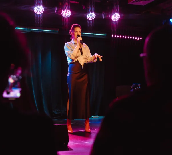 Zdjęcie przedstawia osobę stojącą na scenie i mówiącą do mikrofonu podczas występu. Scena jest oświetlona kolorowymi reflektorami, a na pierwszym planie widoczne są sylwetki publiczności.