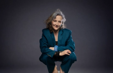 zdjęcie przedstawia artystę. Kobieta kuca na taborecie, jest ubrana w zielony garnitur i ma gołe stopy