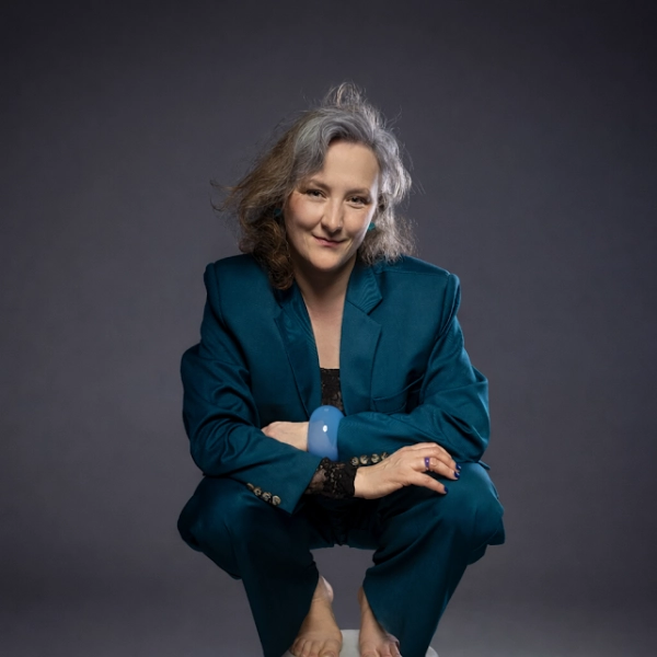 zdjęcie przedstawia artystę. Kobieta kuca na taborecie, jest ubrana w zielony garnitur i ma gołe stopy