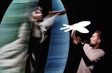 Artysta wykonuje dynamiczną akrobację w kole cyrkowym (Cyr wheel), tworząc świetlne smugi na ciemnej scenie.