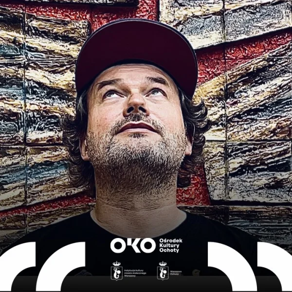 Mężczyzna z brodą, w czarnej koszulce z kolorowymi ilustracjami i czerwonej czapce z daszkiem, stoi przed dużą, fakturowaną instalacją artystyczną w odcieniach brązu, czerwieni i zieleni. Patrzy w górę, jakby zamyślony lub zaintrygowany.