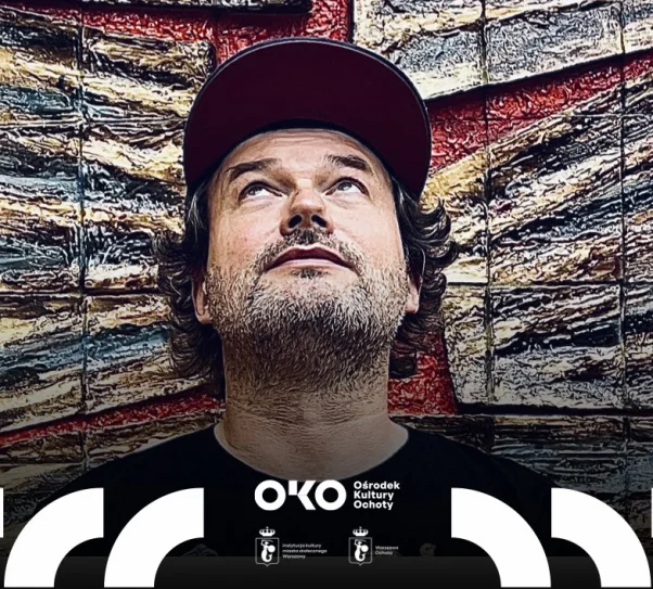 Mężczyzna z brodą, w czarnej koszulce z kolorowymi ilustracjami i czerwonej czapce z daszkiem, stoi przed dużą, fakturowaną instalacją artystyczną w odcieniach brązu, czerwieni i zieleni. Patrzy w górę, jakby zamyślony lub zaintrygowany.