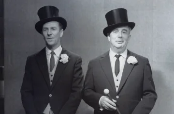 Two elegantly dressed men stand side by side against a plain background. Both wear tailcoats, vests, ties, and top hats, each with a white flower pinned to the lapel. The man on the right is holding a cane with a rounded handle. They look toward the camera.