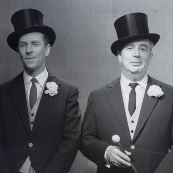 Dwóch elegancko ubranych mężczyzn stoi obok siebie na neutralnym tle. Obydwaj mają na sobie fraki, kamizelki, krawaty i cylindry, a w butonierkach białe kwiaty. Mężczyzna po prawej trzyma laskę zakończoną kulą. Patrzą w kierunku obiektywu.
