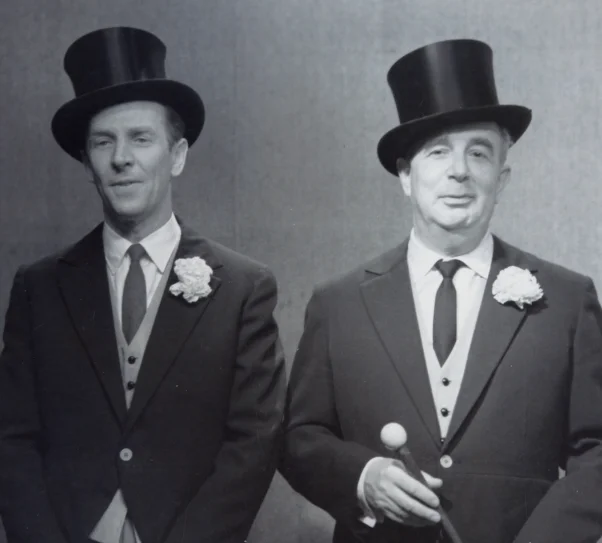 Dwóch elegancko ubranych mężczyzn stoi obok siebie na neutralnym tle. Obydwaj mają na sobie fraki, kamizelki, krawaty i cylindry, a w butonierkach białe kwiaty. Mężczyzna po prawej trzyma laskę zakończoną kulą. Patrzą w kierunku obiektywu.
