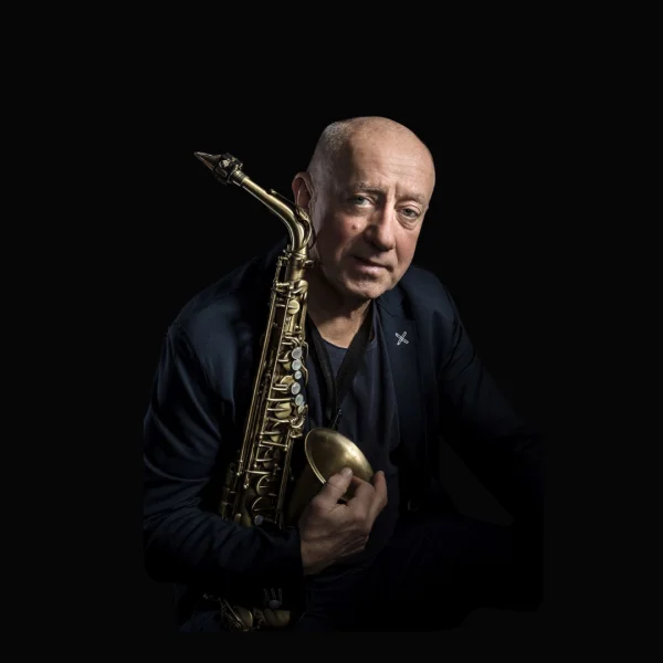 Portret starszego mężczyzny trzymającego saksofon. Siedzi w półmroku, oświetlony miękkim światłem z boku. Ma krótkie, siwiejące włosy i nosi ciemny T-shirt oraz granatową marynarkę. Patrzy spokojnie w stronę aparatu, a instrument opiera na ramieniu, obejmując go dłonią.