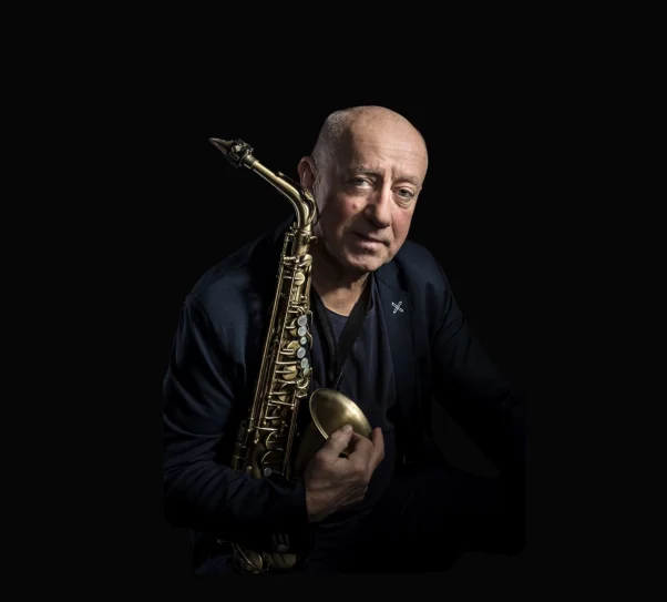 Portret starszego mężczyzny trzymającego saksofon. Siedzi w półmroku, oświetlony miękkim światłem z boku. Ma krótkie, siwiejące włosy i nosi ciemny T-shirt oraz granatową marynarkę. Patrzy spokojnie w stronę aparatu, a instrument opiera na ramieniu, obejmując go dłonią.