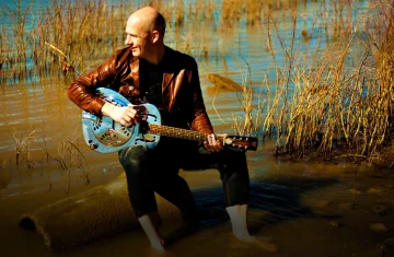 Mężczyzna łysy z brązowej kurtce siedzi na pieńku w wodzie i trzyma niebieską gitarę.