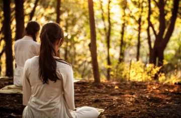 Na zdjęciu widać osoby siedzące w lesie w pozycji medytacyjnej, ubrane w białe stroje. Siedzą na matach, odwrócone plecami do obiektywu, w otoczeniu drzew i miękkiego światła zachodzącego słońca, które tworzy ciepłą, spokojną atmosferę.