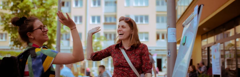 Dwie kobiety przybijają sobie piątkę. Uśmiechają się do siebie. Kobieta po lewej ma ciemne włosy i kolorową koszulę, a kobieta po prawej ma jasne włosy i pomarańczową koszulę. Nad nimi widać kawałek baneru z napisem "Dzień Sąsiada". W tle jest blok mieszkalny.