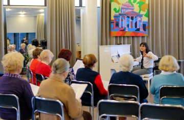 grupa seniorów siedzi na krzesłach, przed nimi siedzi instruktorka i przygrywa im na pianine.