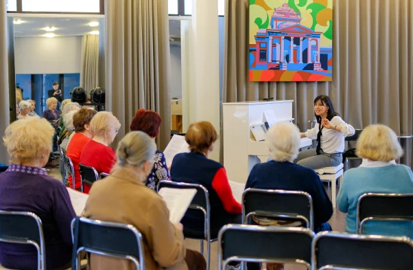 grupa seniorów siedzi na krzesłach, przed nimi siedzi instruktorka i przygrywa im na pianine.