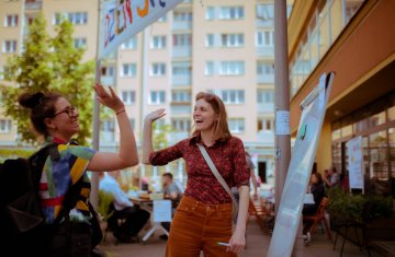 Dwie kobiety przybijają sobie piątkę. Uśmiechają się do siebie. Kobieta po lewej ma ciemne włosy i kolorową koszulę, a kobieta po prawej ma jasne włosy i pomarańczową koszulę. Nad nimi widać kawałek baneru z napisem "Dzień Sąsiada". W tle jest blok mieszkalny.