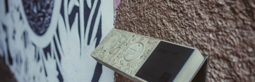 Zbliżenie na tyflografikę zamontowaną na chropowatej ścianie budynku; wypukłe elementy są wyczuwalne dotykiem, w tle fragment czarno-białego muralu o organicznych kształtach.